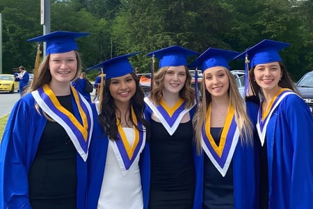 Cinco alumnas con vestidos de graduación azules y amarillos están juntas al aire libre en el terreno de la Howe Sound Secondary School.