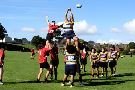 Un grupo de estudiantes juega rugby en el campo deportivo de Howick College frente a edificios y árboles.