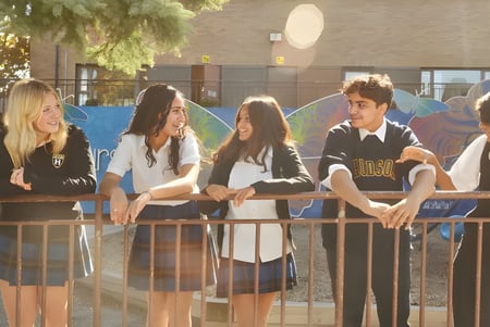 Un grupo de estudiantes está sentado junto en un banco en el exterior del Hudson College con árboles y edificios de fondo.