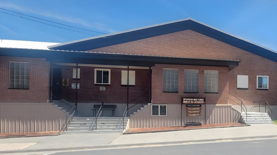 El edificio de ladrillo de dos pisos de Huntington High School con techo inclinado y escalera de entrada en una calle con líneas eléctricas.