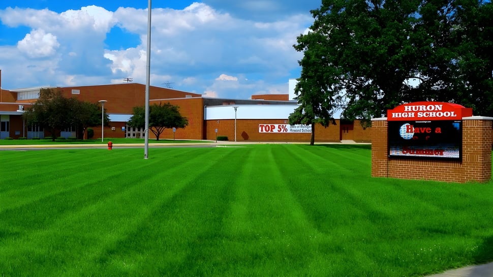 El edificio de la Huron High School se encuentra detrás de un cuidado césped verde bajo un cielo azul con nubes blancas.
