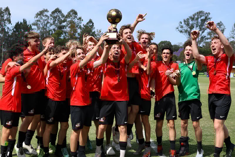 Un grupo de jóvenes futbolistas masculinos en camisetas rojas de Hurtwood House celebra junto con un trofeo en un campo de césped.