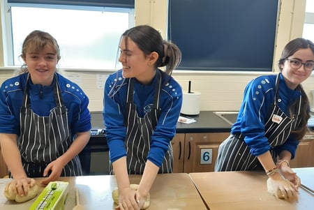Tres estudiantes del Iascaigh College están de pie en uniformes azules junto a una mesa en el aula con una pizarra al fondo.