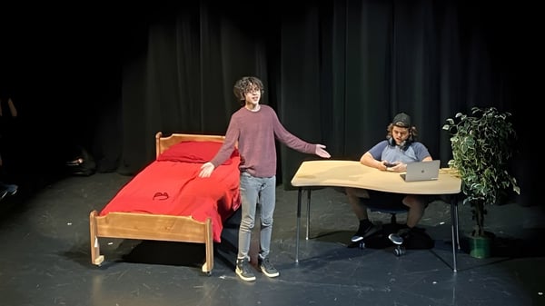 Una estudiante con camiseta rosa está en el escenario de la Imagine High Integrated Arts and Technology Secondary School con un sofá rojo y una persona en la mesa al fondo.