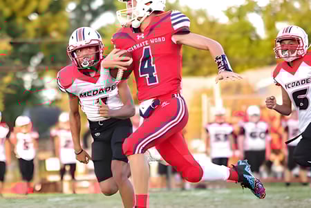 Estudiantes de la Incarnate Word Academy juegan al fútbol en el campo deportivo con espectadores al fondo.