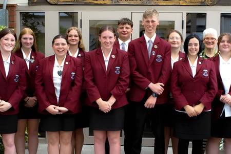 Un grupo de estudiantes en uniformes rojos está frente a un edificio en el campus de Inglewood High School con un vehículo militar de fondo.