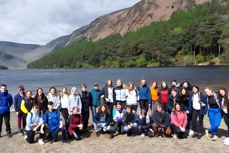 Un grupo de estudiantes del Inver College se reúne en una playa frente a un paisaje montañoso con un lago al fondo.