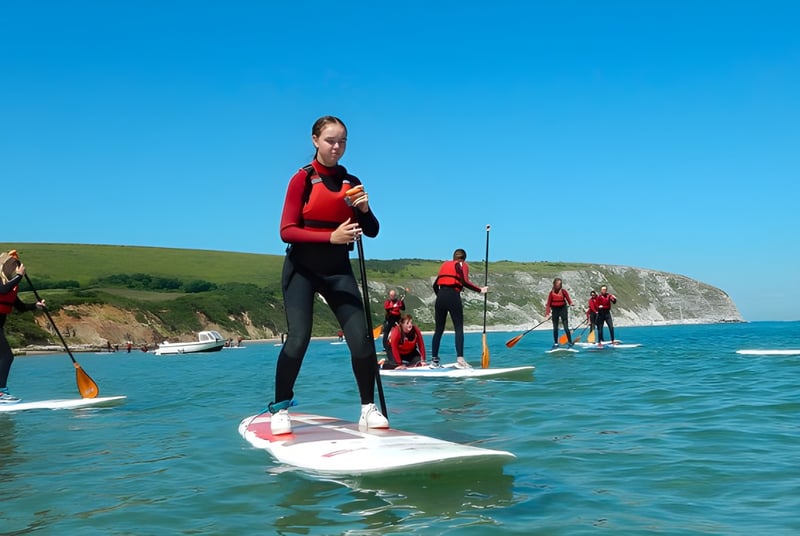 Estudiantes de Ipswich High School están de pie sobre paddleboards en un cuerpo de agua con un paisaje costero de fondo.