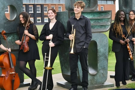 Un grupo de músicos sostiene instrumentos frente a un edificio de ladrillo en el terreno de la Ipswich School.