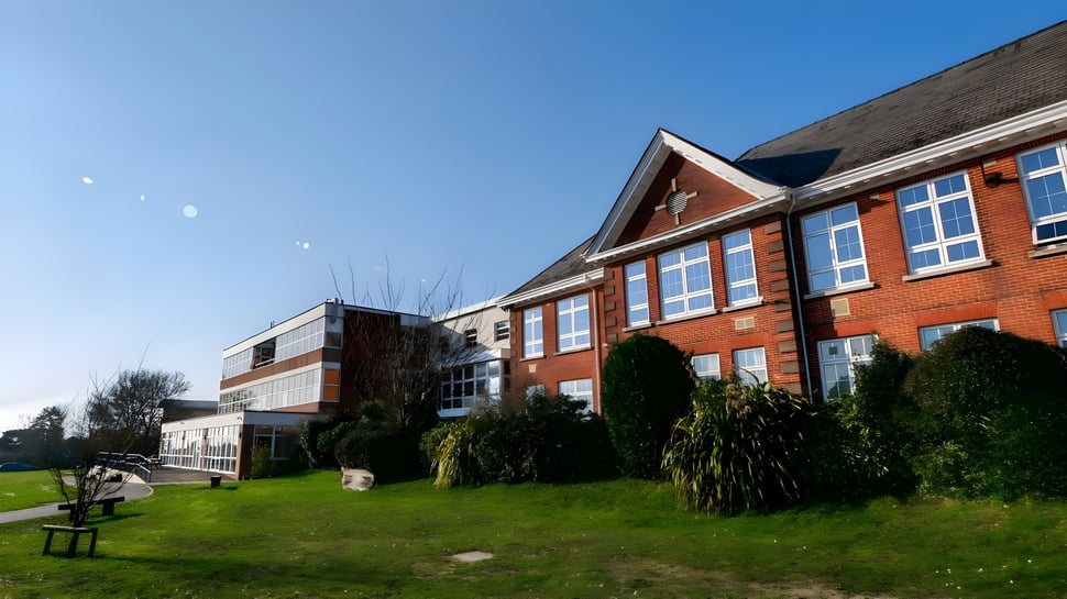 El edificio de ladrillo de varios pisos del Itchen Sixth Form College con una gran fachada de ventanas frente a una superficie de césped y árboles se puede ver bajo un cielo despejado.