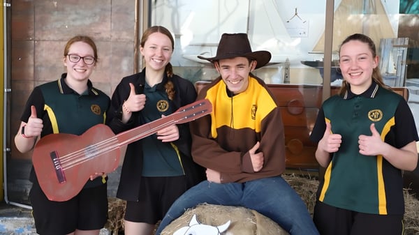 Un grupo de estudiantes de la James Nash State High School está con instrumentos musicales frente a un edificio de madera.