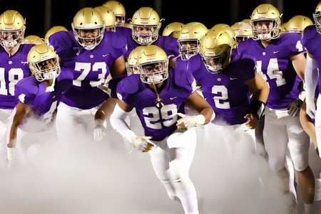 Un grupo de futbolistas en uniformes morados está de pie por la tarde en el campo de la John Bapst Memorial High School.