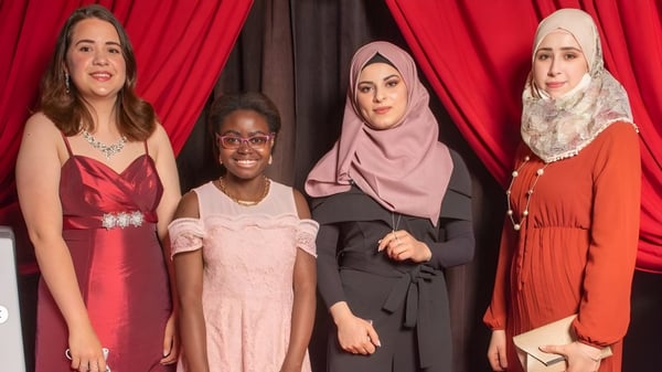 Cuatro estudiantes en vestidos coloridos y hijabs están de pie frente a una cortina roja en el terreno de la John Barsby Secondary School.