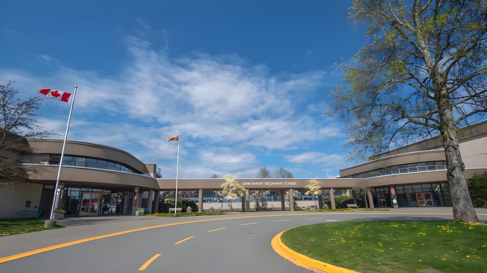 El moderno edificio de la John Barsby Secondary School está rodeado de árboles y muestra una bandera canadiense frente a un cielo azul.
