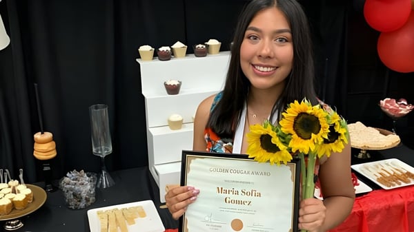 Una alumna de la John F. Kennedy High School sostiene un certificado y girasoles frente a una mesa con pasteles y decoraciones.