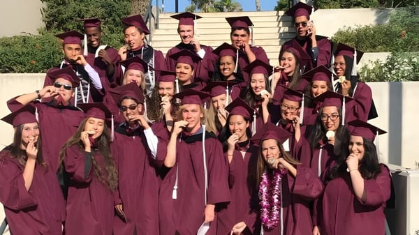Graduadas y graduados de la John F. Kennedy High School en túnicas burdeos están frente a un edificio.