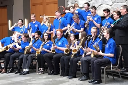 Estudiantes de la John Maland High School en uniformes azules durante un ensayo de orquesta frente a un edificio de piedra.