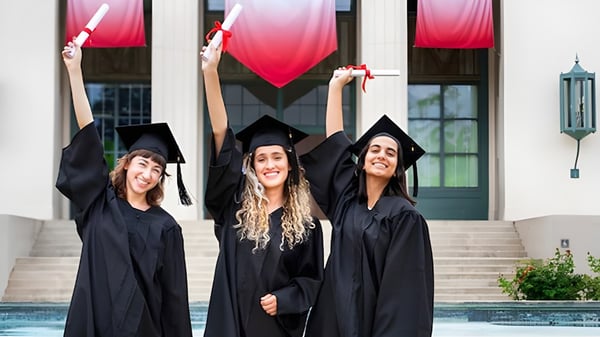 Tres estudiantes de la John Oliver Secondary School están juntas en ropa de graduación y sostienen orgullosamente sus diplomas frente al edificio escolar con piscina.