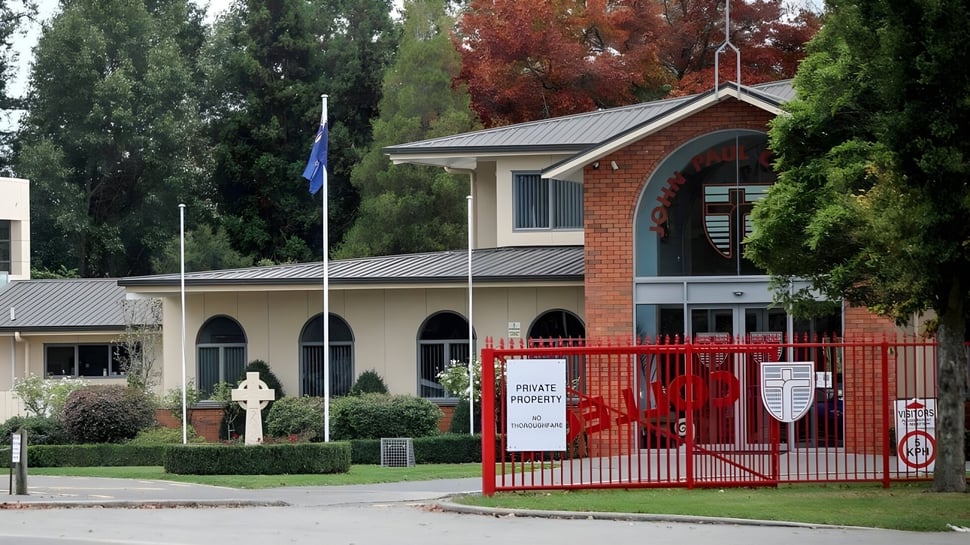 El edificio de ladrillo con puerta roja del John Paul College está rodeado de árboles y áreas verdes.