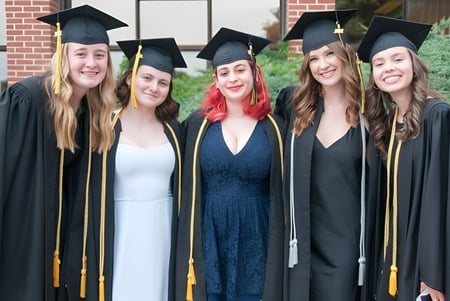 Cinco graduadas de la John Rennie High School están frente a un edificio de ladrillo celebrando su graduación.