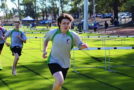 Una persona con ropa deportiva blanca corre en el campo de deportes de la Kalamunda Senior High School superando obstáculos en un campo de césped.