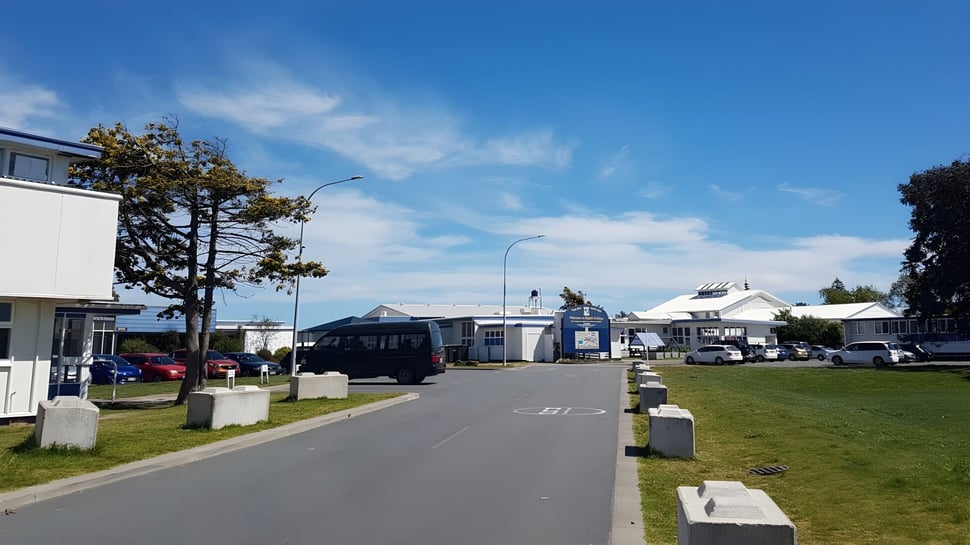 Un camino pavimentado lleva a través de un área verde en el campus del Kapiti College con varios edificios al fondo.