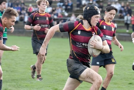 Un grupo de jugadores de rugby del Kapiti College juega un intenso partido en un campo de hierba con espectadores al fondo.