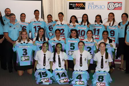 Un grupo de alumnas de la Keebra Park State High School está en uniformes deportivos frente a un panel Públicoitario.