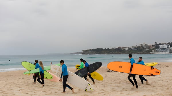 Un grupo de alumnas y alumnos de la Keebra Park State High School lleva coloridas tablas de surf en la playa con la ciudad de fondo.