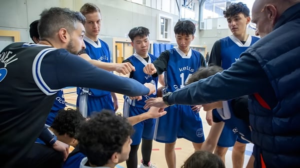 Un grupo de jóvenes jugadores de baloncesto en camisetas azules escucha las instrucciones del entrenador en el terreno de Kells Academy.