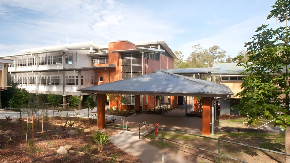 El moderno edificio de varios pisos de la Kenmore State High School con fachada de vidrio y área exterior cubierta rodeada de árboles.