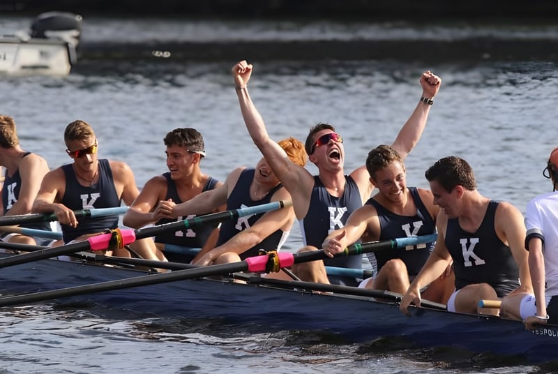 Un grupo de remeros en un bote celebra en el agua frente a la vista de la ciudad de la Kent School.