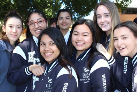 Un grupo de alumnas del Kiara College está en uniforme deportivo juntas al aire libre frente a árboles.