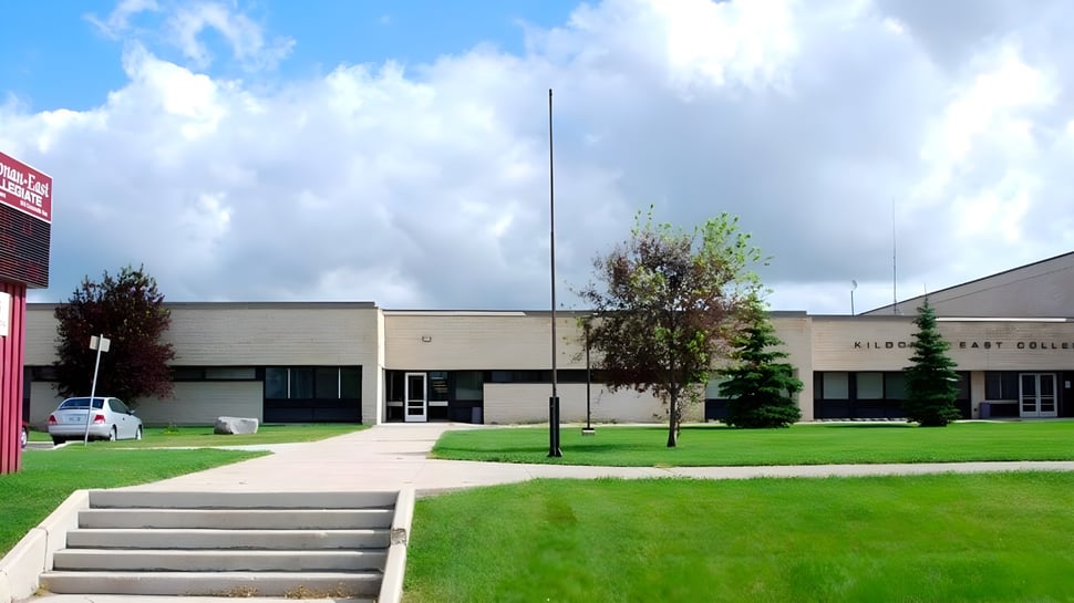 El moderno edificio escolar de varios pisos de Kildonan East Collegiate con una superficie de césped y árboles en primer plano bajo un cielo nublado.