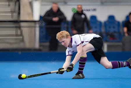 Una persona juega al hockey sobre césped en el campo de césped artificial azul de la Kimbolton School frente a un grupo de espectadores.