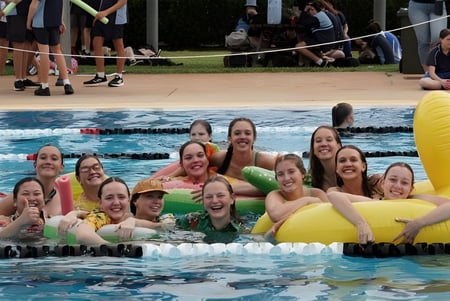 Un grupo de alumnas de la Kingaroy State High School se reúne en una piscina frente a espectadores y una red de voleibol.