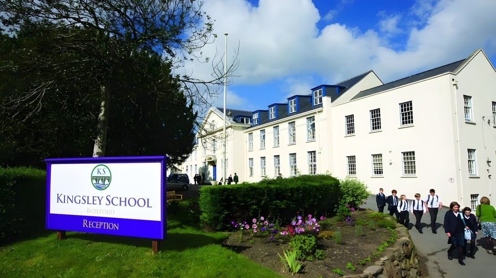 El edificio blanco de varios pisos de la Kingsley School está rodeado de árboles y muestra un camino con personas al fondo.
