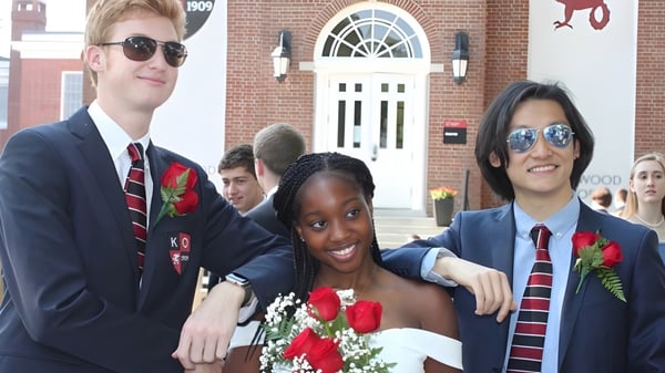 Tres estudiantes de la Kingswood Oxford School posan con ropa formal frente a un edificio de ladrillo con entrada arqueada.