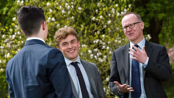 Tres hombres en trajes mantienen una conversación frente al fondo verde con árboles en el terreno de la Kingswood School.