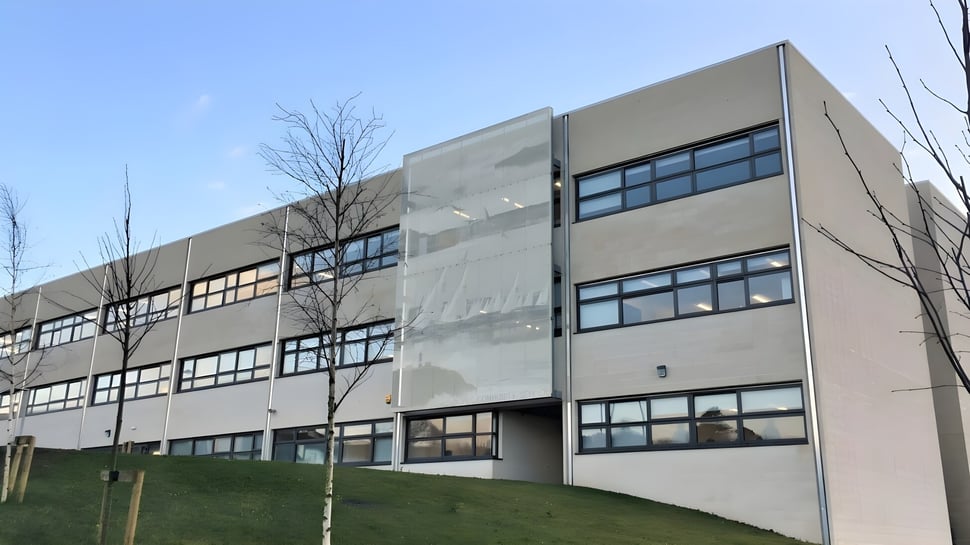 El moderno edificio de varios pisos de la Kinsale Community School se encuentra en una ladera cubierta de hierba con árboles desnudos al fondo.