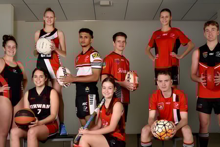 Un grupo de jóvenes atletas de la Kirwan State High School posan en el gimnasio con uniformes de equipo y equipos deportivos.