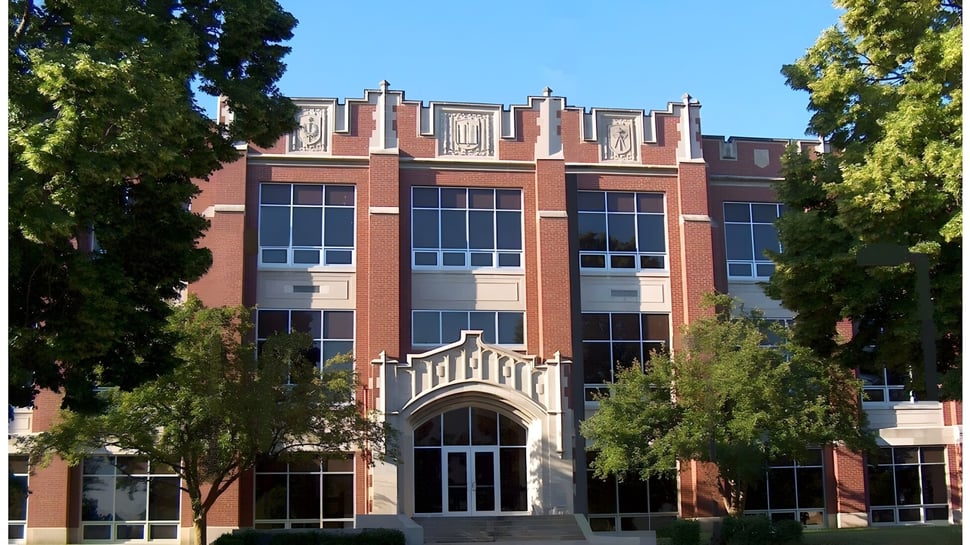 El gran edificio de ladrillo con ventanas de arco de la Kokomo High School está rodeado de árboles y áreas verdes.