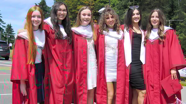 Un grupo de estudiantes de la Kwalikum Secondary School está de pie en túnicas de graduación rojas frente a un fondo boscoso.