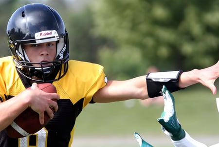 Un jugador de fútbol americano de La Salle Secondary School está con casco y balón en un campo de césped frente a árboles.