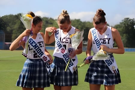 Tres alumnas de Lacey Township High School en uniformes de animadoras con pompones están en un campo con árboles y cielo azul.
