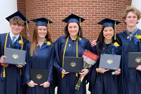 Un grupo de cinco graduados de la Lake Ridge Academy está frente a una pared de ladrillo sosteniendo sus diplomas.