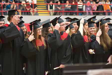 Un grupo de graduadas y graduados del Lambton Central Collegiate & Vocational School está de pie en togas negras frente a una pared colorida con banderas.