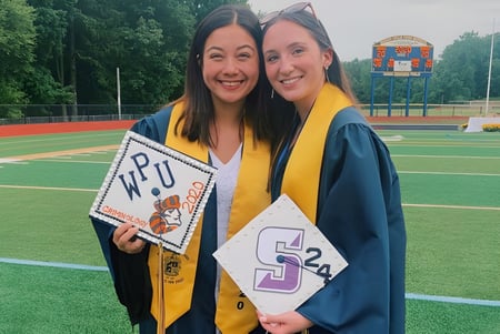 Dos alumnas del Las Virgenes Unified School District en ropa de graduación están en un campo deportivo sosteniendo carteles de graduación.