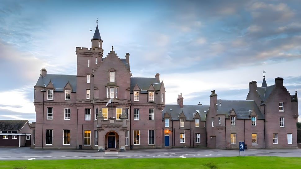 El magnífico edificio parecido a un castillo de la Lathallan School con torres se encuentra frente a edificios más pequeños y un prado bajo un cielo nublado.