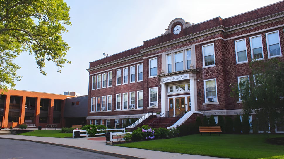 El edificio de ladrillo con torre del reloj en el campus de la Lawrence Woodmere Academy está rodeado de árboles verdes y un camino pavimentado.